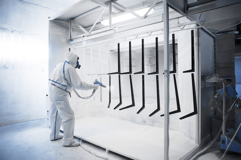 Trabalhador usando equipamentos de proteção individual realiza a pintura eletrostática a pó de peças metálicas em uma cabine industrial especializada.